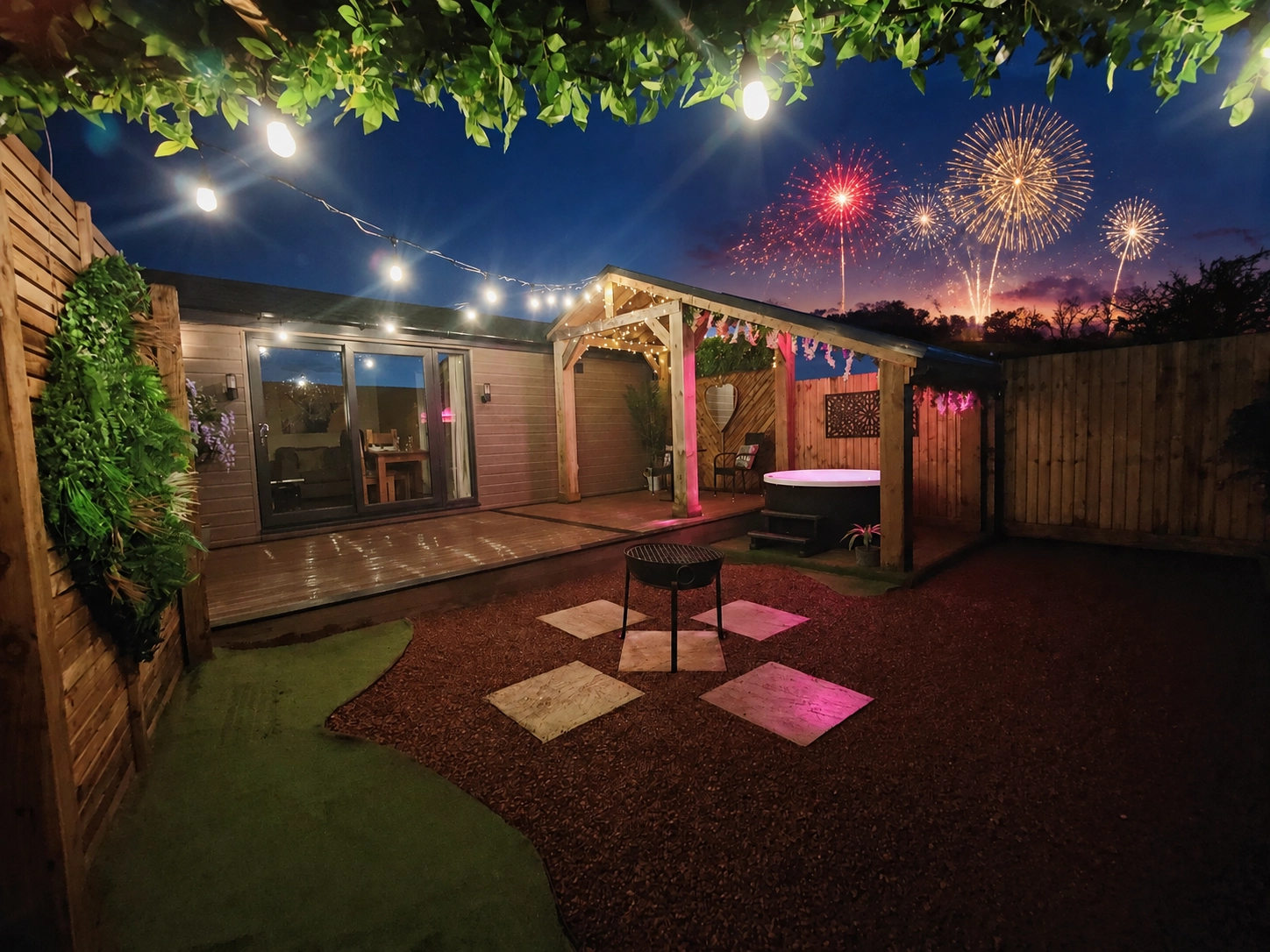 Lodge patio at twilight with an undercover hot tub, fire pit and fireworks bursting in the night sky